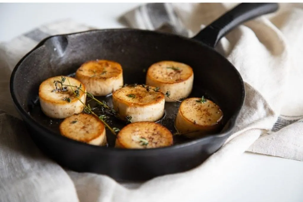 Sliced, browned medallions of king trumpet/king oyster mushrooms in a black cast iron skillet, garnished with fresh thyme, on a light cloth background.