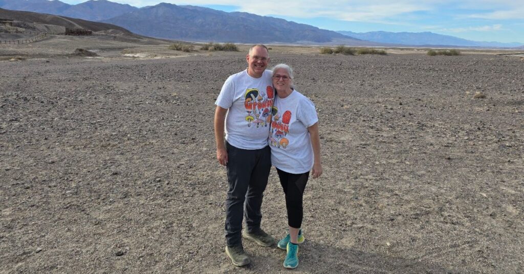 A smiling couple stands close together on a rocky desert landscape, wearing matching graphic mushroom T-shirts. Mountains and a clear blue sky are in the background.